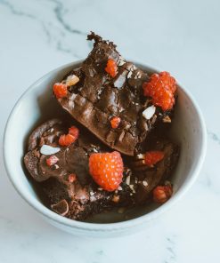 A bowl filled with rich chocolate brownies topped with fresh raspberries and nuts on a white marble surface.